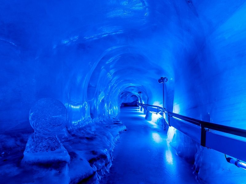 A tunnel carved into glacier ice, lit blue, with a handrail along its wall