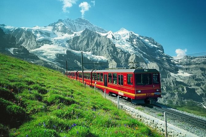 A red and cream cogwheel train on an alpine meadow below a glaciated massif